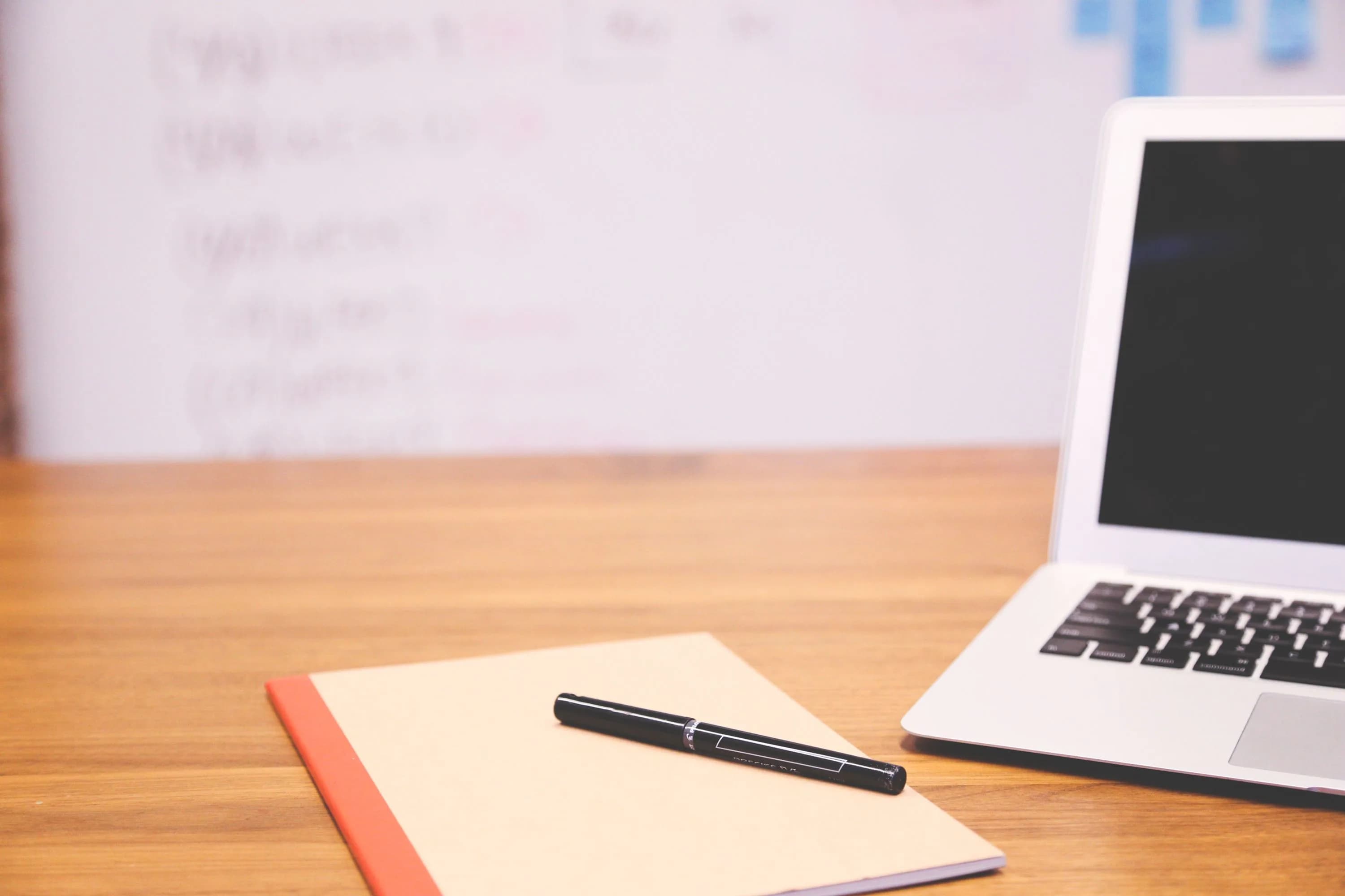 Workspace with a laptop, notebook and pen on a wooden desk with a blurred whiteboard behind