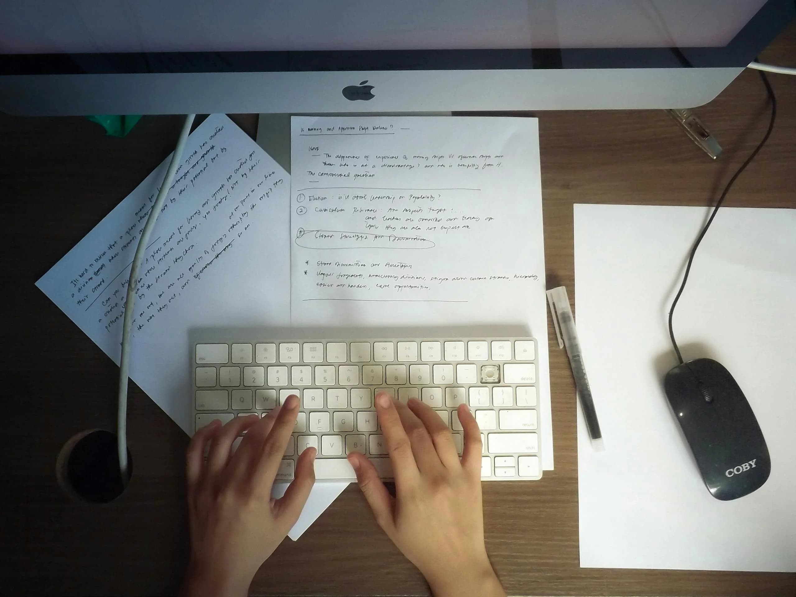 Hands on a keyboard beside paper notes on a wooden desk, the slow path that voice-to-text shortens