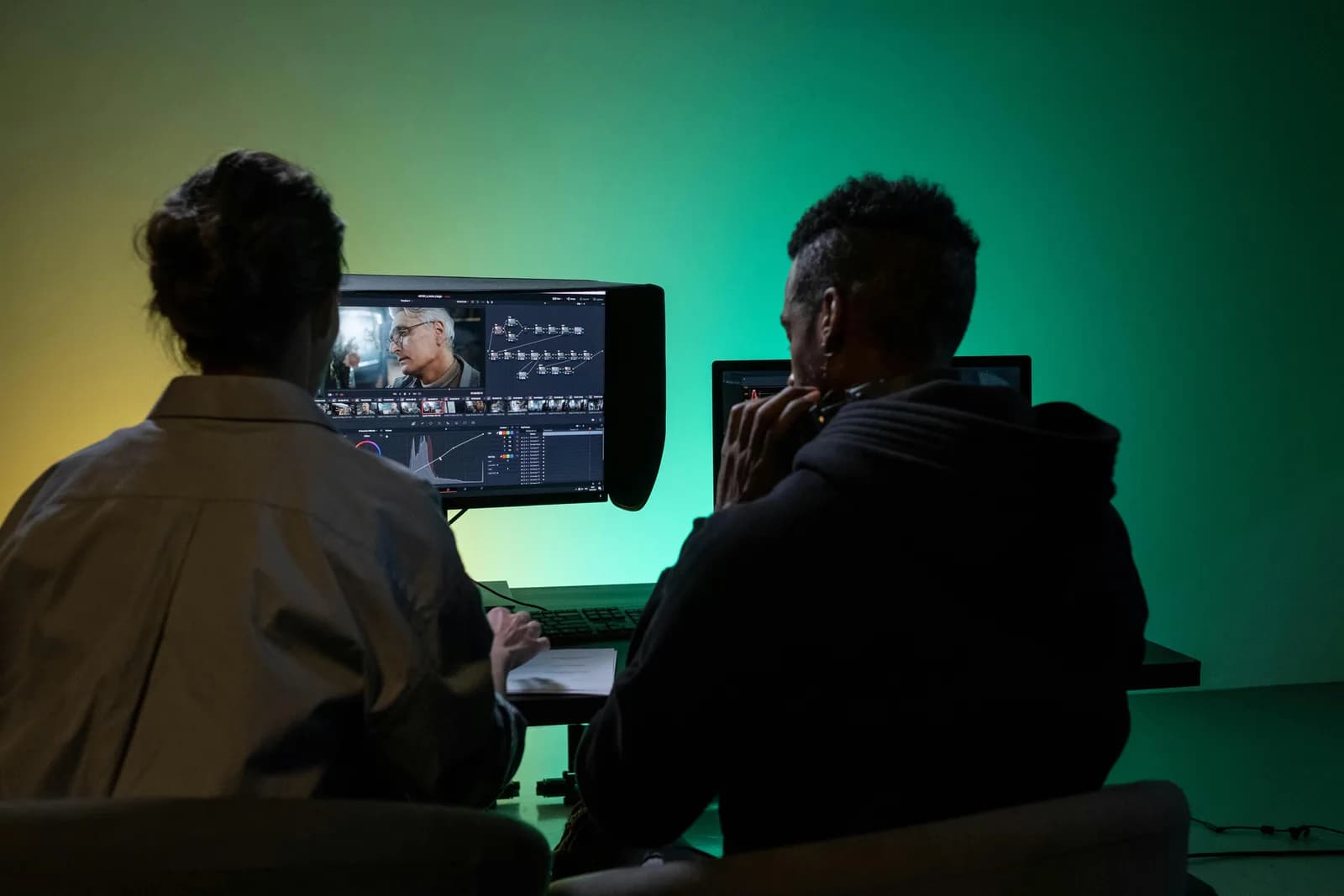 Back view of two colleagues editing footage on a dual-monitor setup, a team review workflow