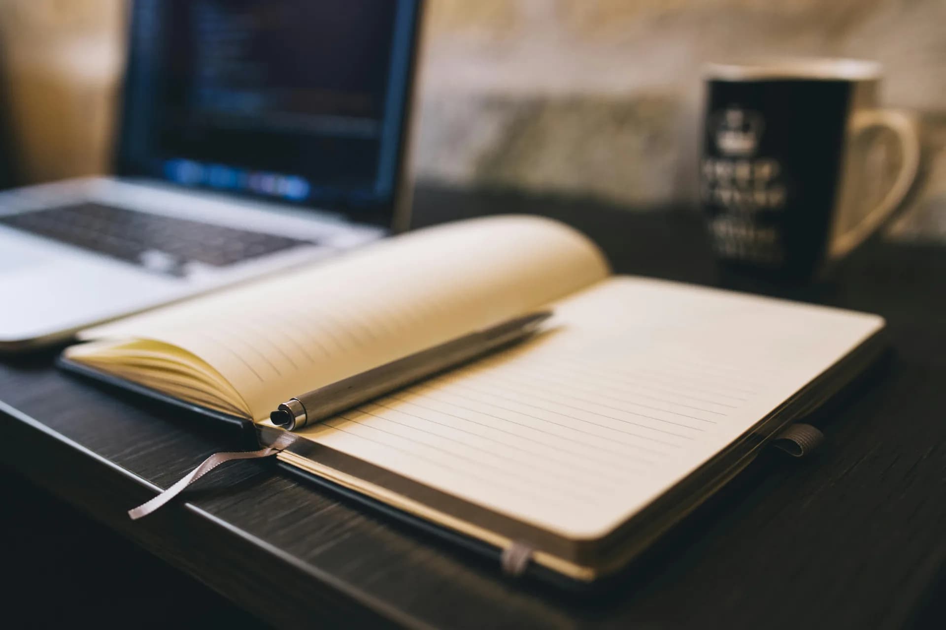 Open notebook and pen beside a laptop on a wooden desk, a note-taking workspace for dictating into Evernote