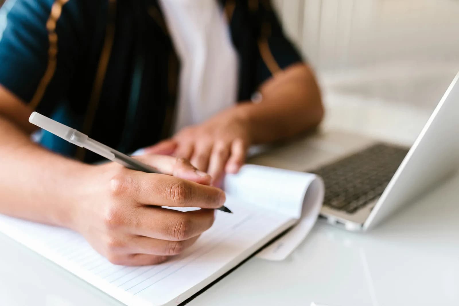 Hands jotting a quick note in a paper notebook beside a laptop, evoking fast idea capture