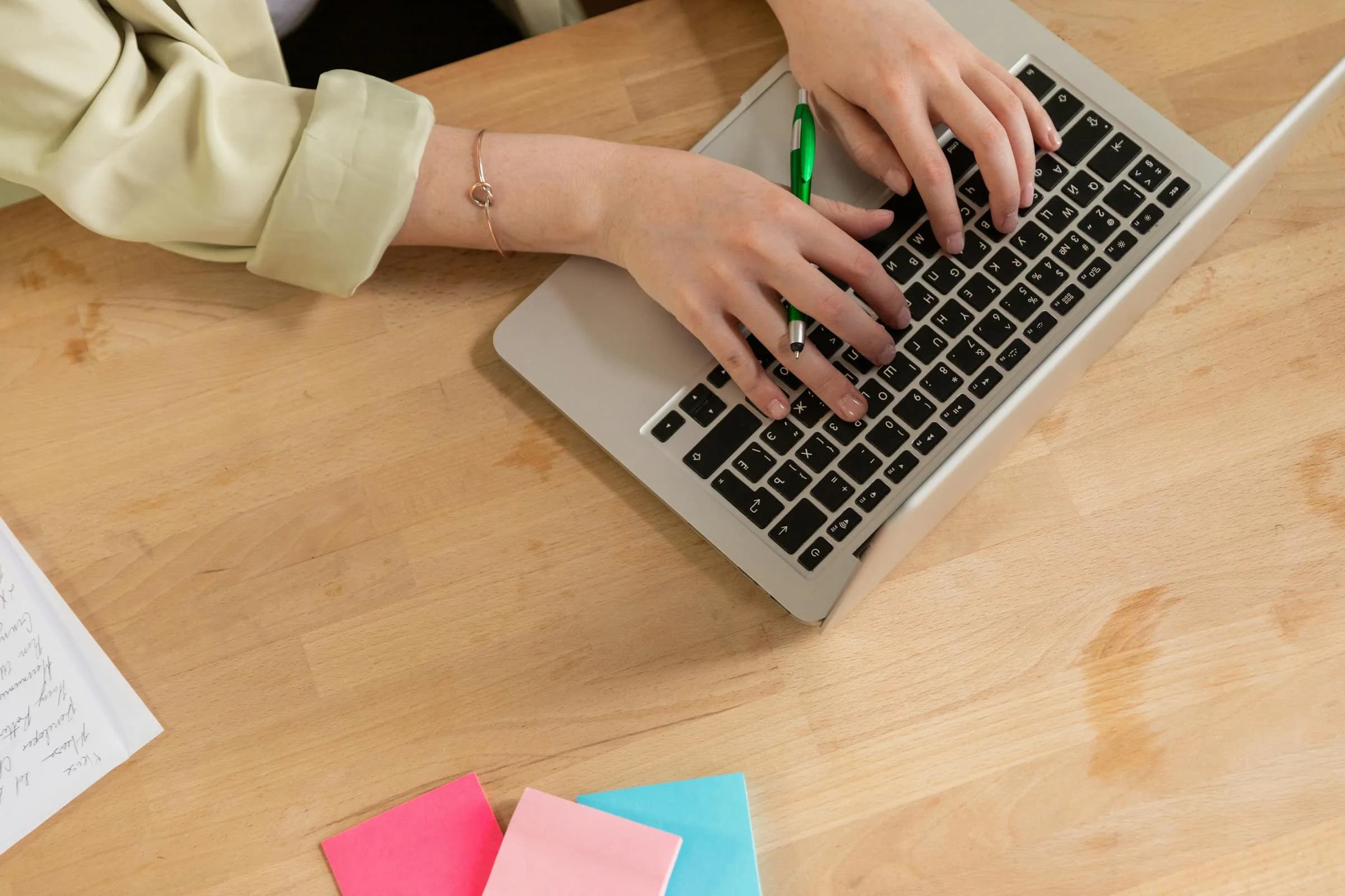 Mani che digitano su un laptop accanto a una penna e a foglietti adesivi colorati, in contrasto tra la digitazione manuale e la dettatura