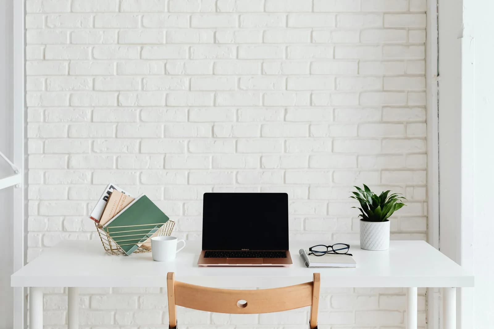 A calm, uncluttered desk with a laptop, a plant, and a couple of books against a white brick wall