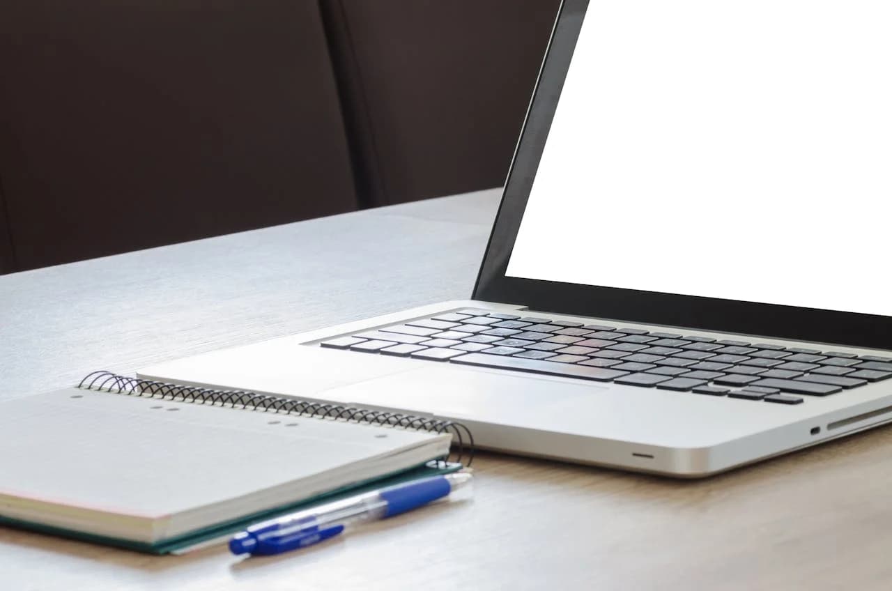 Open laptop next to a notebook and pen on a wooden desk, ready for writing or dictation