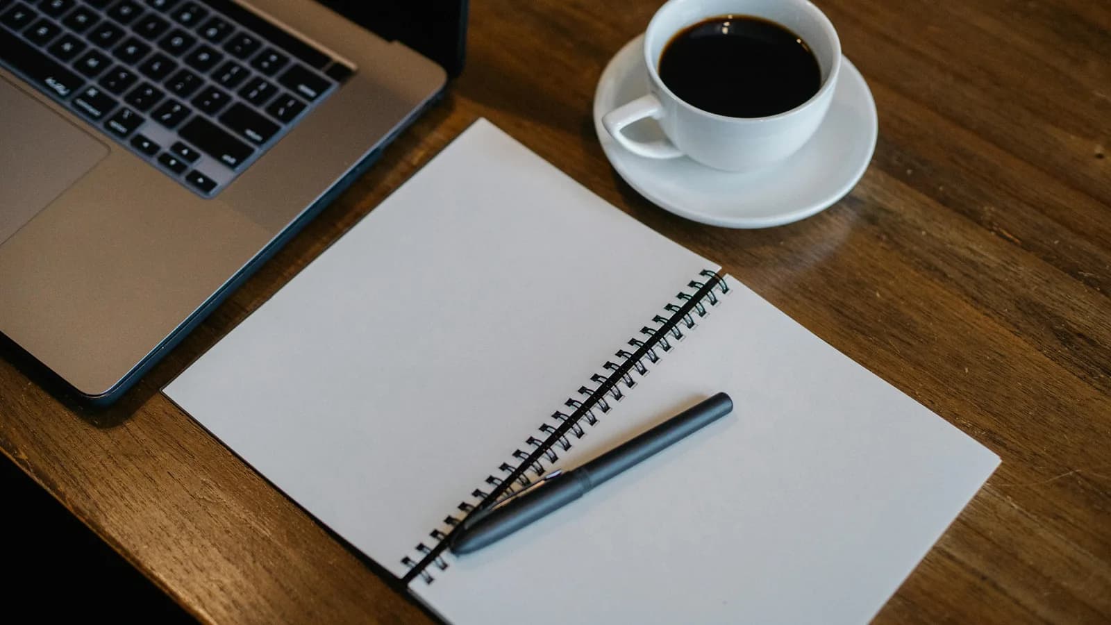 A dark desk with an open laptop and a notebook beside a coffee, evoking note-taking by voice