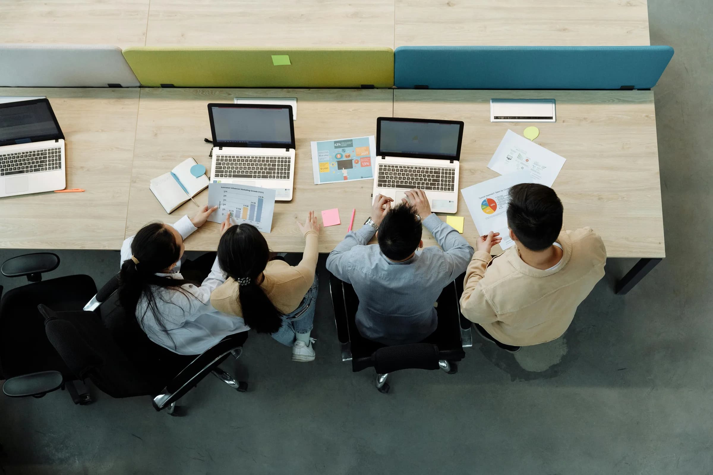 Aerial view of four colleagues working on laptops at a shared desk, evoking team chat and collaboration